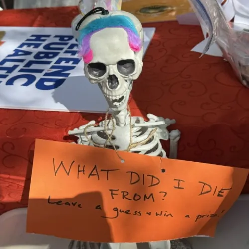 A skeleton holding a sign reading, "What did I die from?" Behind it is a Defend Public Health sign