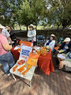 Table at an outdoor event, with a Defend Public Health sign and decorated with flowers and pumpkins. Volunteers speak to someone visiting the table.