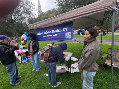 Volunteers talking to people at an outdoor booth with  a sign reading "Defend Public Health -- CT"