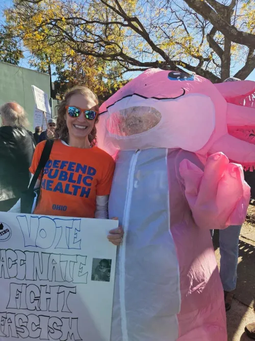 Woman in a Defend Public Health shirt holding a sign, next to a pink inflatable cartoon character